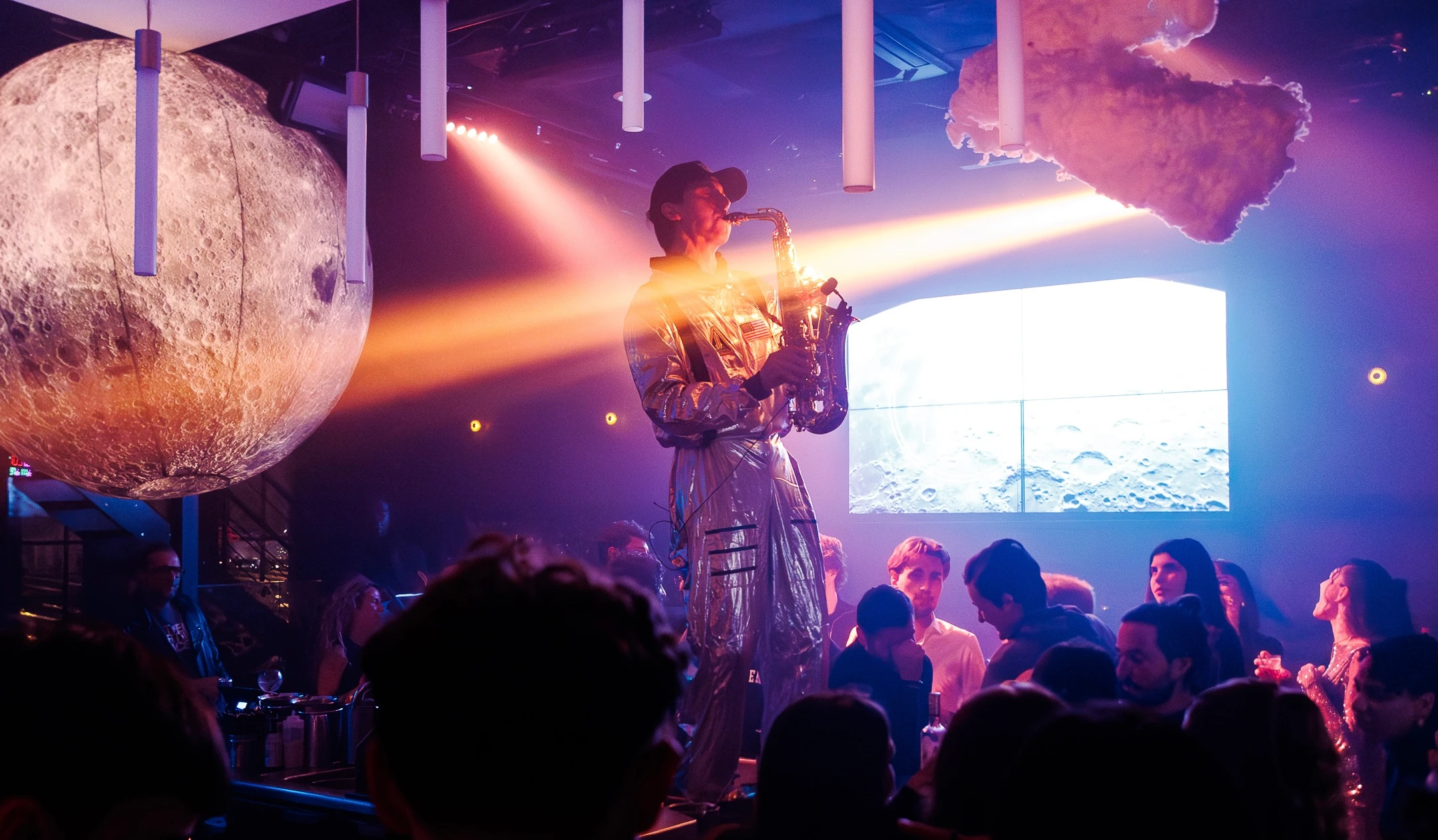 Performer en combinaison futuriste jouant du saxophone sur une scène de club.