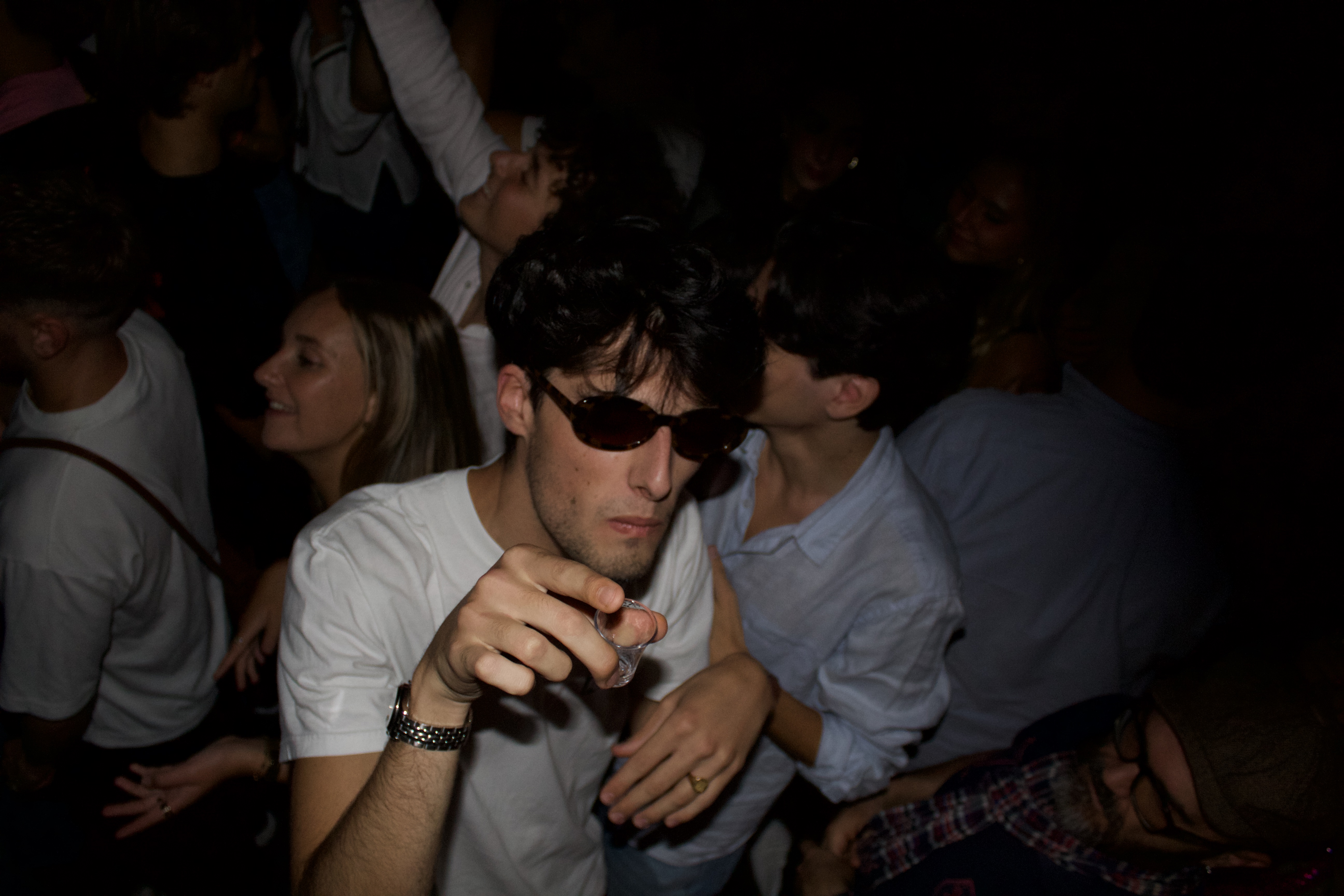Jeune homme vetu de lunette de soleil pointe la camera avec un shot à la main