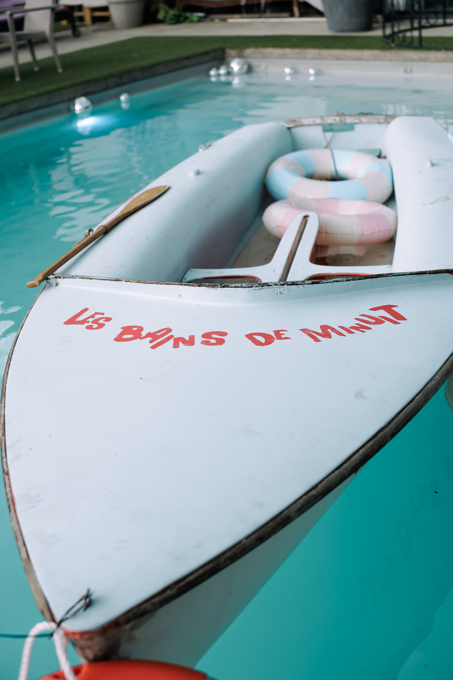 Bateau blanc avec une pagaie et deux bouées pastel, flottant dans une piscine, avec l'inscription 'Les Bains de Minuit' sur la proue.