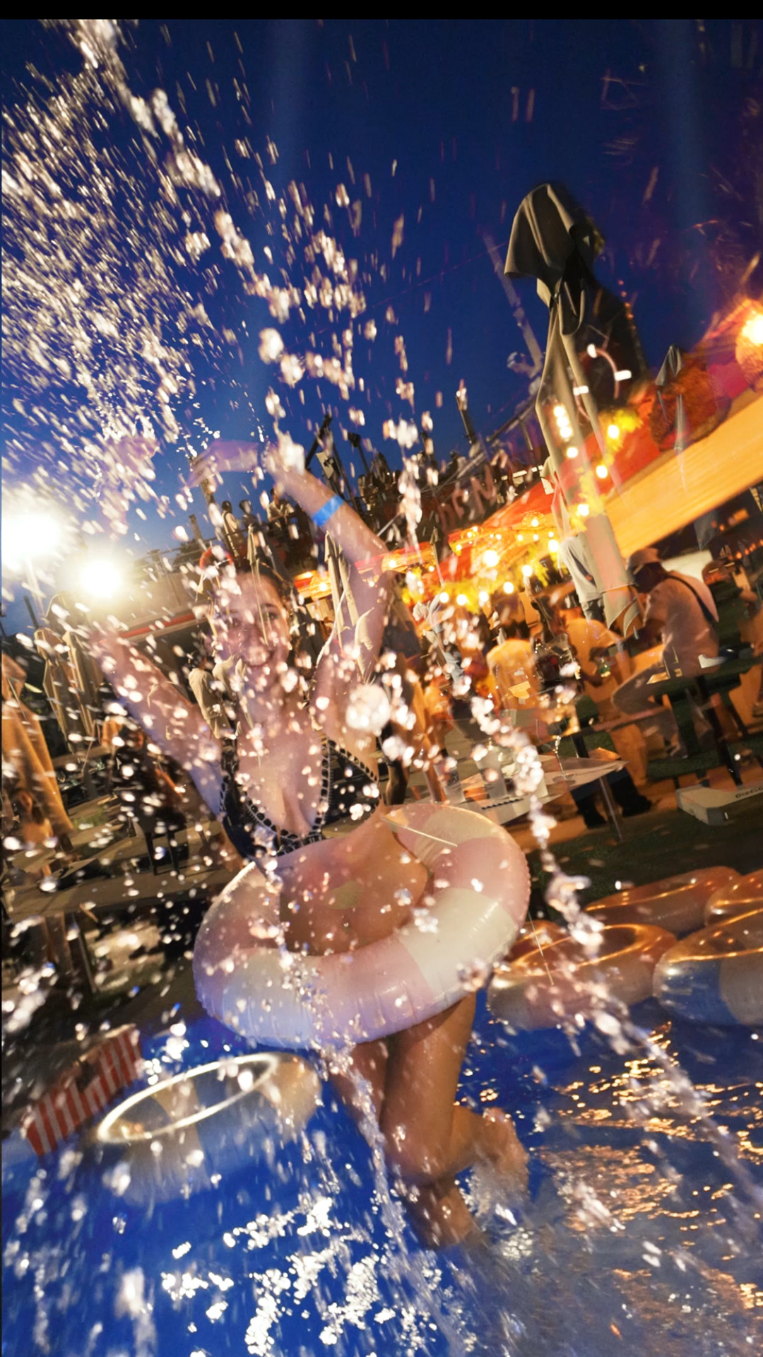 Femme dans une bouée, éclaboussée par l’eau sous les lumières festives du soir, ambiance joyeuse d’une pool party.