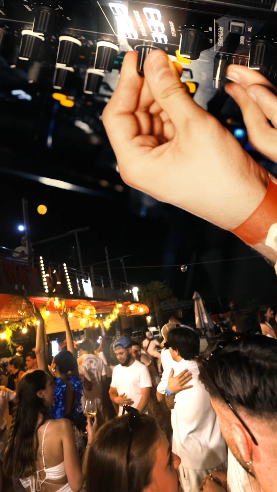 Main sur une table de mixage et foule habillée en blanc dans une ambiance estivale animée par des guirlandes lumineuses.