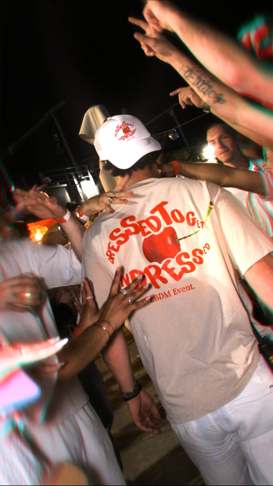 Personne de dos avec un t-shirt 'Dressed to get undressed', entourée de mains lors d’une soirée festive nocturne.