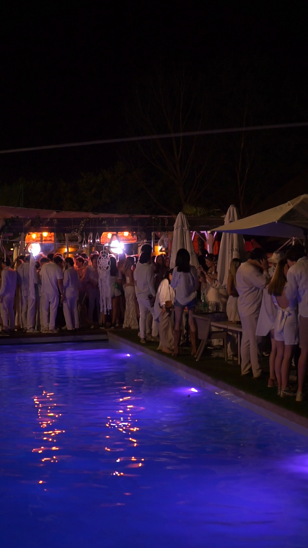 Soirée pool party élégante de nuit, éclairée par des lumières violettes dans la piscine, avec des invités en tenue blanche rassemblés sous les parasols.