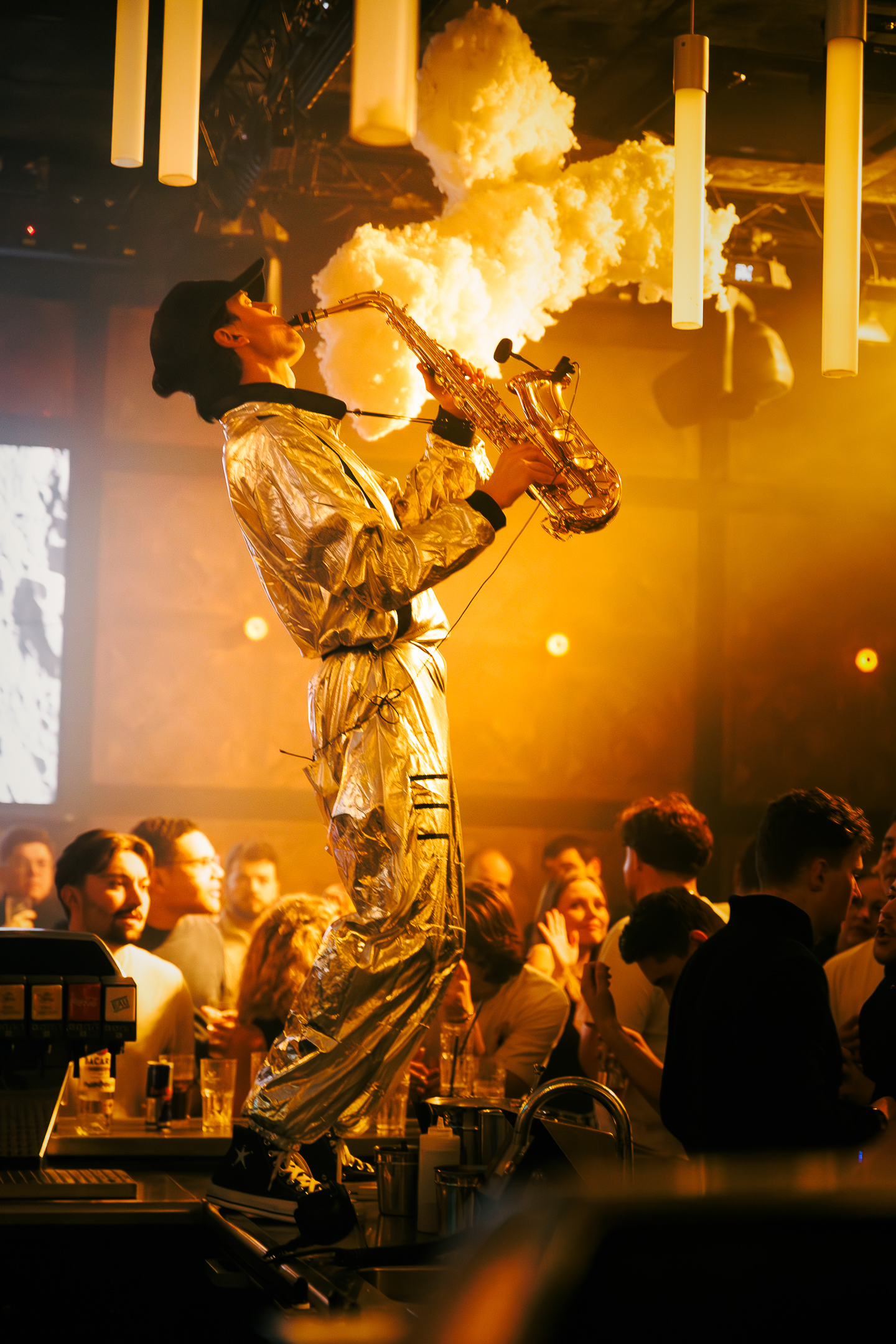 Un saxophoniste en combinaison argentée joue debout sur un comptoir de bar, entouré de spectateurs dans une ambiance jaune et festive.