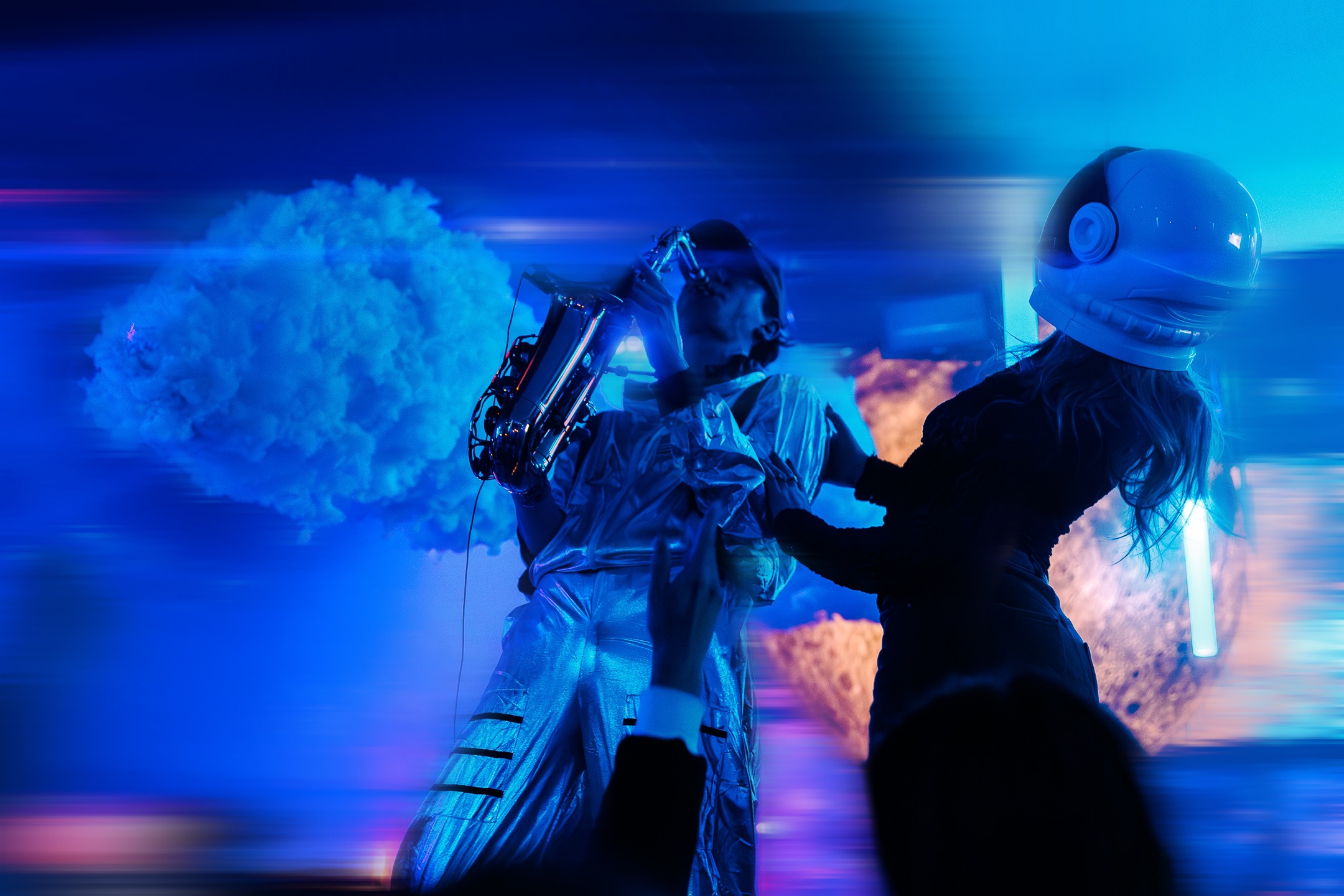 Un musicien en combinaison spatiale joue du saxophone dans une ambiance futuriste bleue, accompagné d’une femme portant un casque d’astronaute.