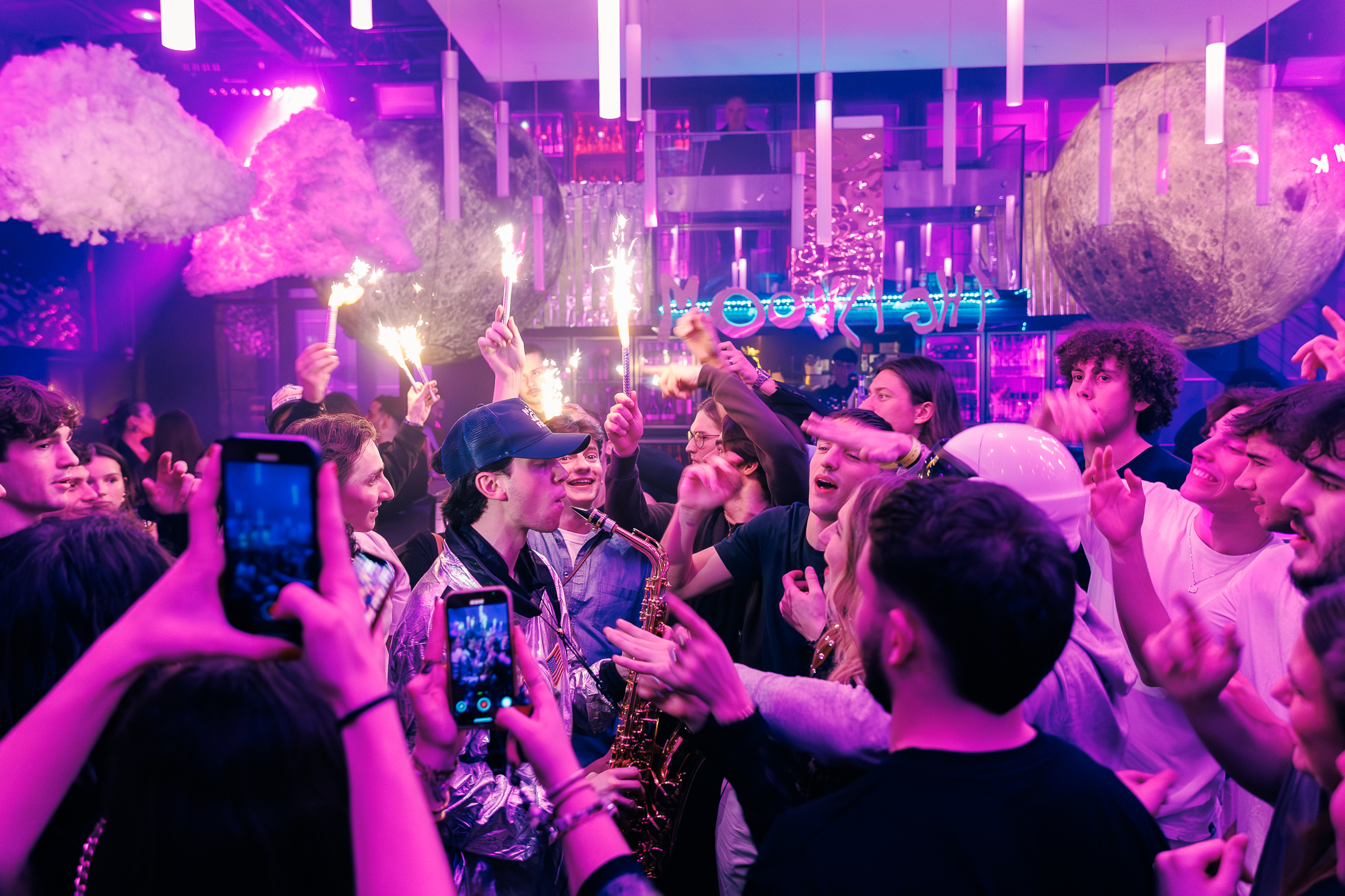 Un saxophoniste au centre d'une foule en liesse dans une boîte de nuit violette, entouré de jeunes brandissant des feux de Bengale et filmant avec leurs téléphones.