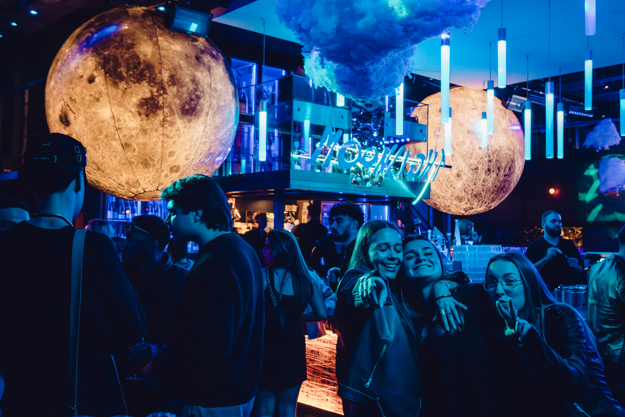 Ambiance de club futuriste avec des jeunes discutant sous des globes lunaires géants et des lumières bleues, dans un décor spatial lumineux.