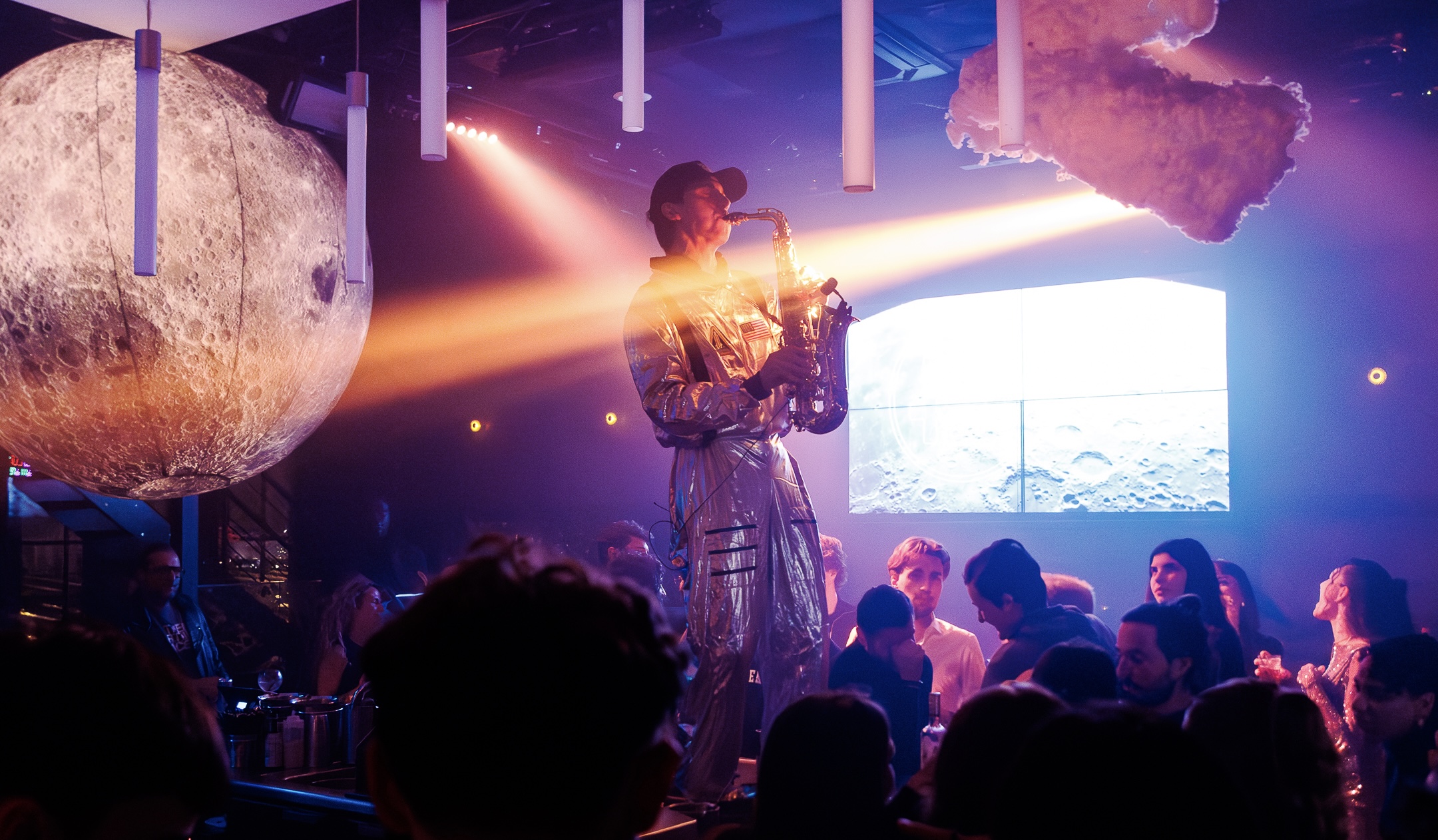 Un saxophoniste en combinaison métallique joue au centre d’un club sous un projecteur orange, devant un écran affichant la surface lunaire et une sphère représentant la Lune.
