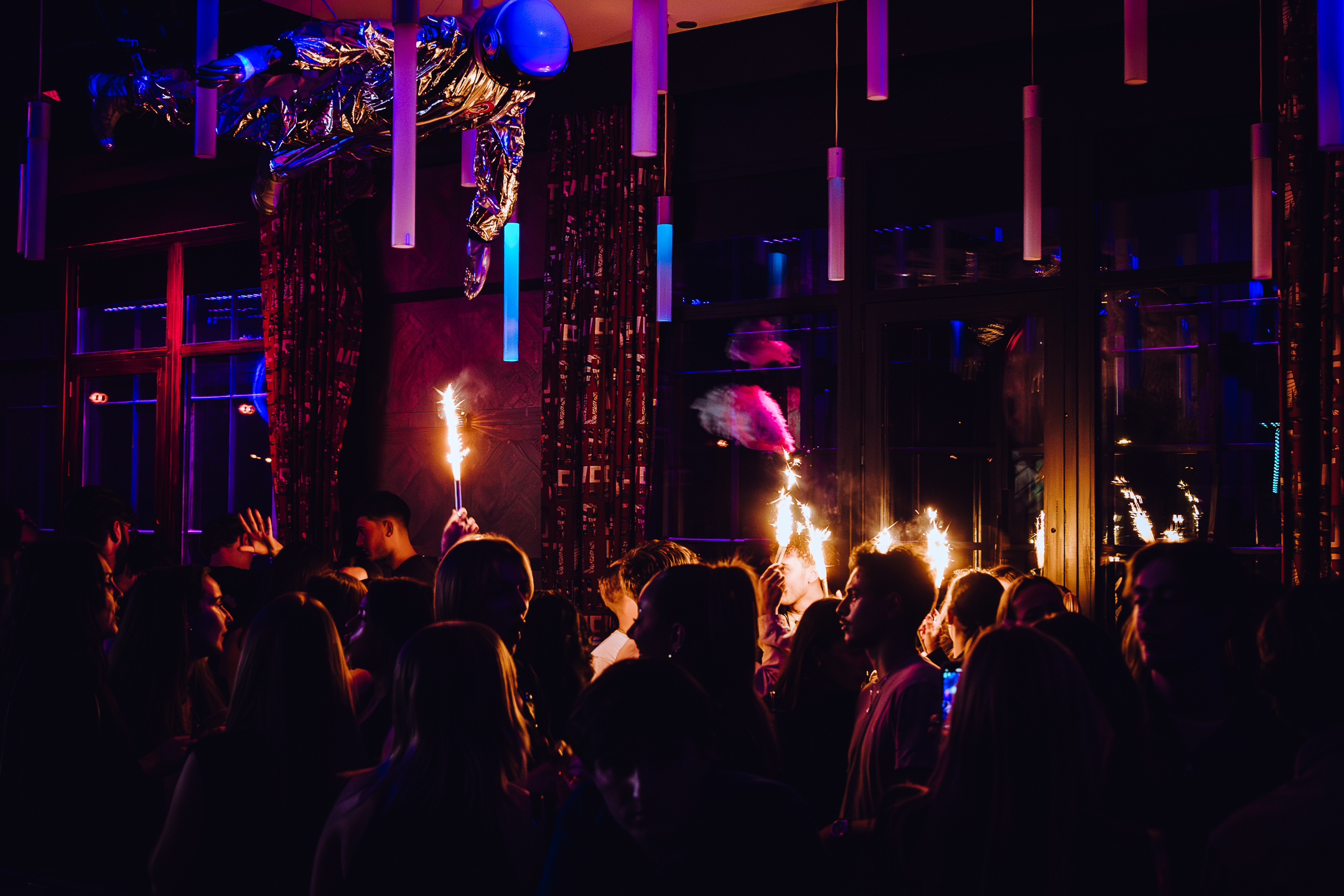 Soirée en club avec une foule dansante et des feux de Bengale, sous une décoration d’astronaute suspendu en combinaison argentée.