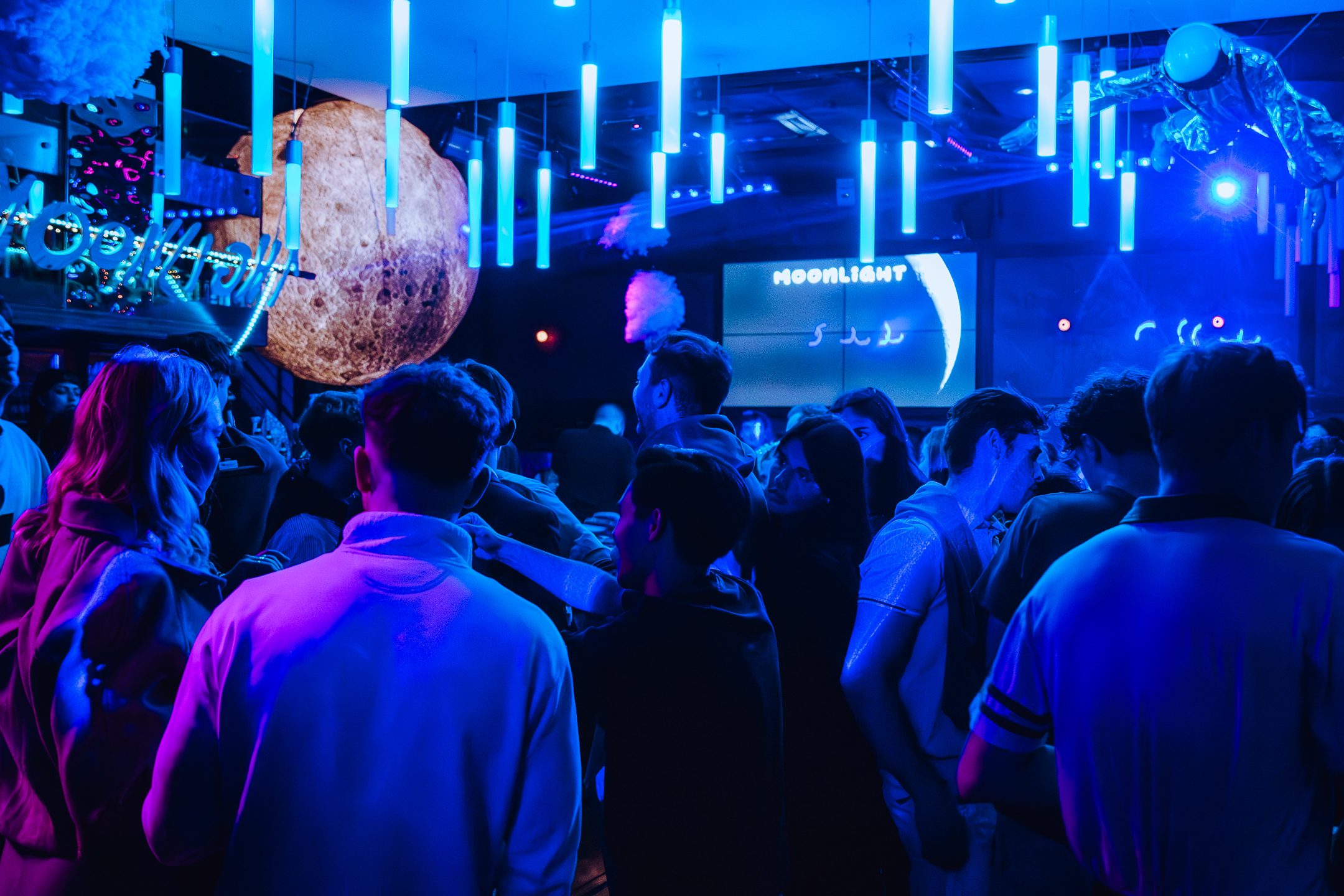 Foule rassemblée dans une boîte de nuit à thème spatial, sous des luminaires bleus et des planètes géantes, avec un écran affichant 'MOONLIGHT'.