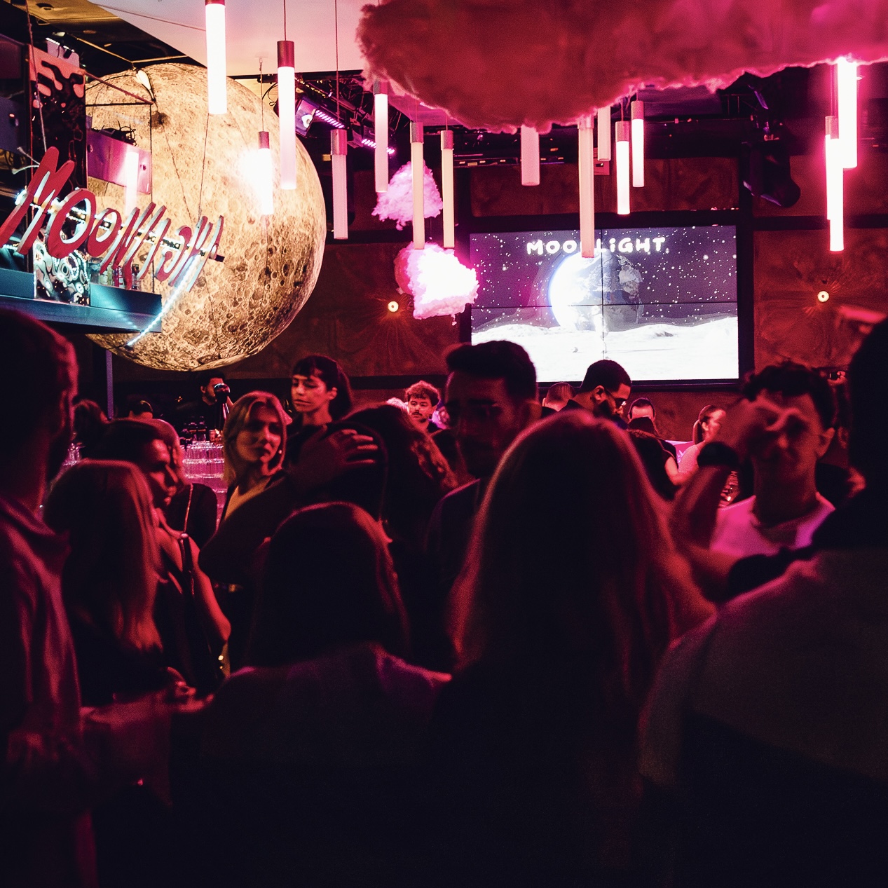Foule réunie dans une boîte de nuit au décor spatial, avec nuages suspendus, grand globe lunaire et écran affichant 'MOONLIGHT' sur fond étoilé.