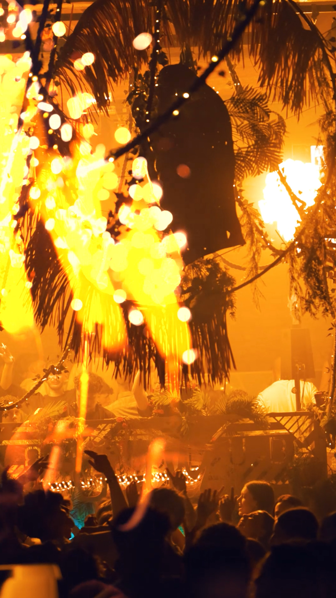 Scène de DJ avec effets lumineux intenses et foule en liesse, ambiance immersive sous une décoration feuillue.