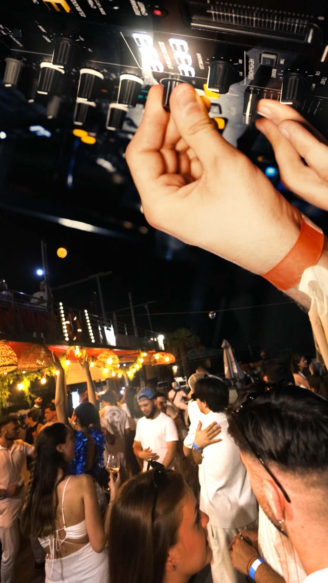 Main sur une table de mixage et foule en plein air, soirée estivale illuminée sous guirlandes et palmiers.