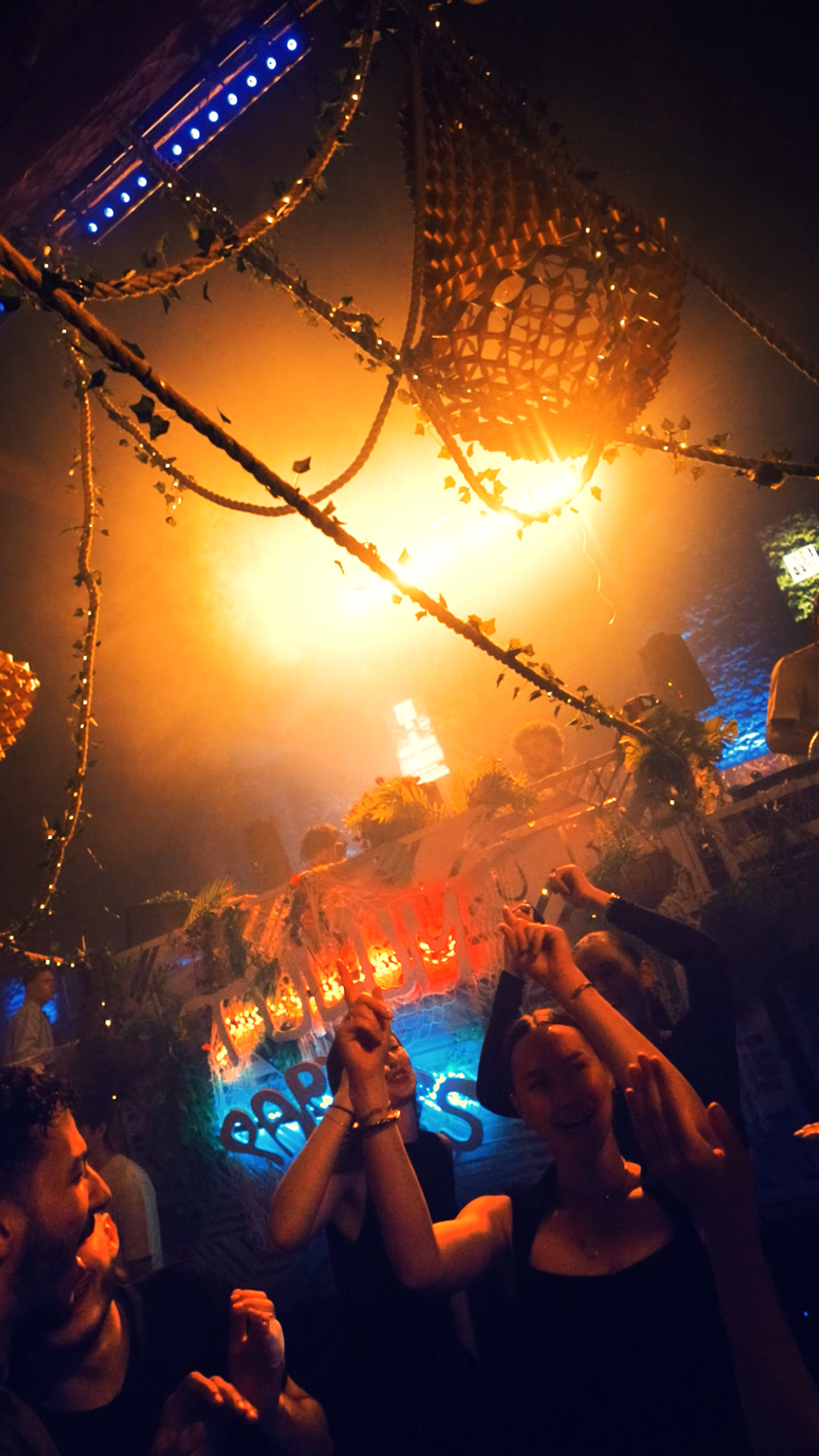 Foule face à la scène, ambiance tamisée et lumière orangée, éléments suspendus dans un décor végétal.